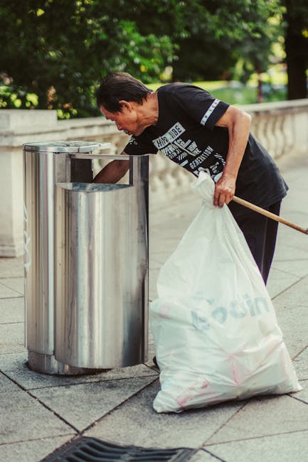 A middle-aged man with dark hair and a focused expression is seen disposing of waste into a large, cylindrical metal bin on a paved outdoor area. He is wearing a black T-shirt with white text and dark trousers. The bin, made of stainless steel, has a rounded top with an opening at the front for depositing rubbish. The surrounding environment includes a stone balustrade in the background and lush green trees suggesting a park. The man is holding a large white plastic bag filled with various waste materials, which he is leaning over the bin to empty. The scene is illuminated by natural daylight, creating clear visibility of the textures of the metal bin, the plastic bag, and the paved surface. This image exemplifies independent rubbish disposal, aligning with the services offered by Rubbish Collection Streatham for efficient waste removal and recycling solutions.
