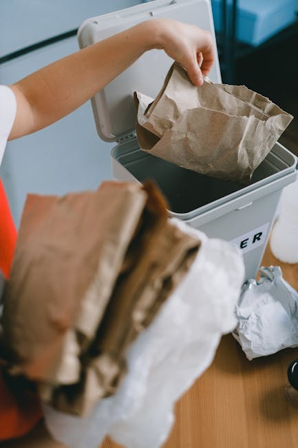Close-up of a person's hand dropping a crumpled brown paper bag into a white rubbish bin with a black interior, positioned on a wooden surface. In the foreground, another crumpled paper bag is held, partly obscuring the view. The background features a kitchen or office environment with a partially visible white appliance or cabinet and some scattered paper waste. The scene emphasizes a waste disposal process, reflecting the practice of independent rubbish collection or on-site clearance, suitable for services like those offered by Rubbish Collection Streatham. The natural lighting highlights the texture of the paper bags and the clean, organized setting, illustrating typical urban waste management involving paper and general refuse sorting.
