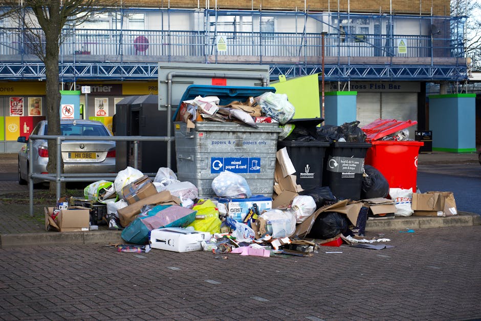 A middle-aged man with dark hair and a focused expression is seen disposing of waste into a large, cylindrical metal bin on a paved outdoor area. He is wearing a black T-shirt with white text and dark trousers. The bin, made of stainless steel, has a rounded top with an opening at the front for depositing rubbish. The surrounding environment includes a stone balustrade in the background and lush green trees suggesting a park. The man is holding a large white plastic bag filled with various waste materials, which he is leaning over the bin to empty. The scene is illuminated by natural daylight, creating clear visibility of the textures of the metal bin, the plastic bag, and the paved surface. This image exemplifies independent rubbish disposal, aligning with the services offered by Rubbish Collection Streatham for efficient waste removal and recycling solutions.