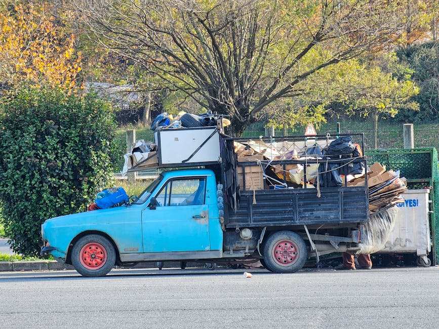 A light blue, small flatbed truck packed with various discarded items is parked along the side of a paved street. The vehicle's body shows signs of wear with faded paint and visible dirt, and the red-painted steel wheels contrast with the truck's blue color. On top of the flatbed are large, irregularly shaped cardboard boxes, black trash bags, and miscellaneous household debris, with some items held in place by ropes. The truck's cab is closed, and a small blue bag rests on the front fender. In the background, a large deciduous tree with yellowing leaves extends its branches over the vehicle, indicating an autumn setting. Behind the tree, a green metal fence runs parallel to the road, and further back, a grassy area with sparse foliage is visible. The scene suggests private waste collection or on-site clearance, typical of independent rubbish removal services like those offered by Rubbish Collection Streatham.