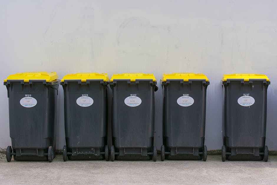 A row of five black wheeled rubbish bins with yellow lids, positioned on a concrete surface against a plain, light-colored wall. Each bin has a white oval label with black text and a barcode, indicating serial or identification numbers. The bins are aligned in a straight line, evenly spaced, with the lids securely closed. The environment appears to be an interior or exterior waste storage area, with no visible surrounding objects or environmental features. The scene is evenly lit, highlighting the texture of the plastic bins and the smooth finish of the wall behind them. The image illustrates a typical setup for private waste collection services, such as those offered by Rubbish Collection Streatham, emphasizing on-site disposal and alternative rubbish management options.
